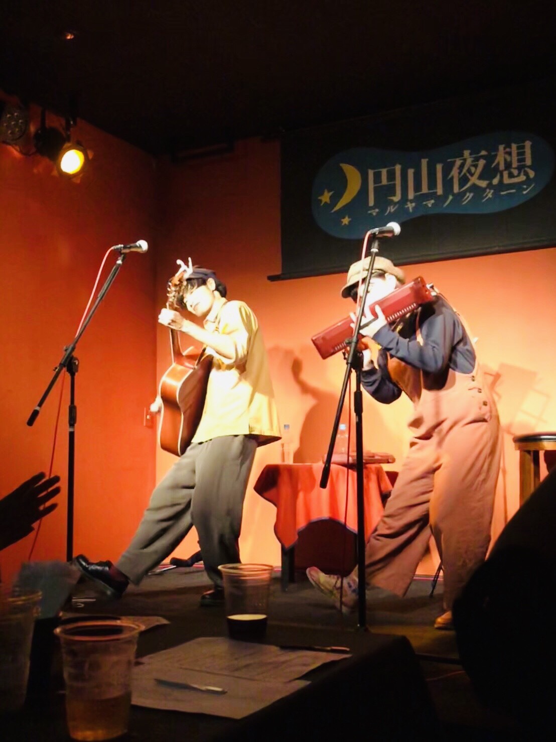 マルヤマ ノクターンのステージでライブを行う「あずけん」の２人。左からアコースティックギターを抱える武内建、ピアニカを演奏する森田杏美。２人で右足を伸ばし、同じポーズを取っているところ。