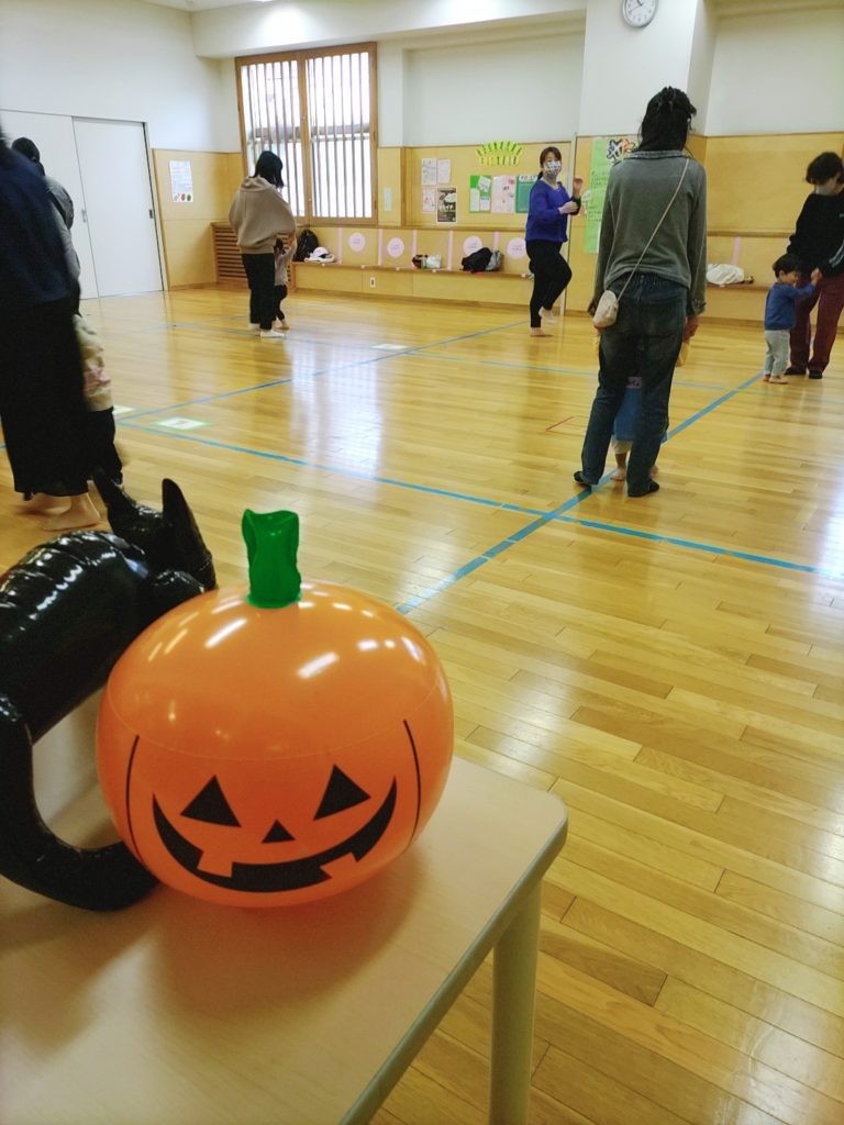 手前にはハロウィンのかぼちゃ、奥には手を繋いでいる数組の親子に呼びかけるリトミック講師